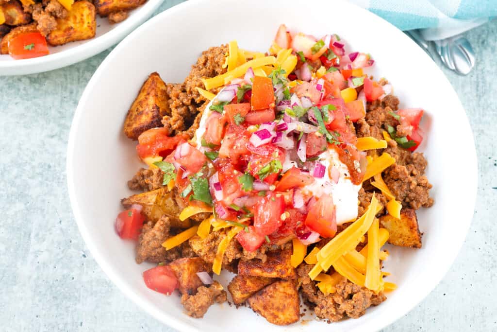 Taco Bowl with Ground beef Taco Meat, Grated Cheddar, Sour Cream, Salsa, and Pico de Gallo on a bed of Taco Potatoes — all in a white low-side serving dish on a concrete countertop.