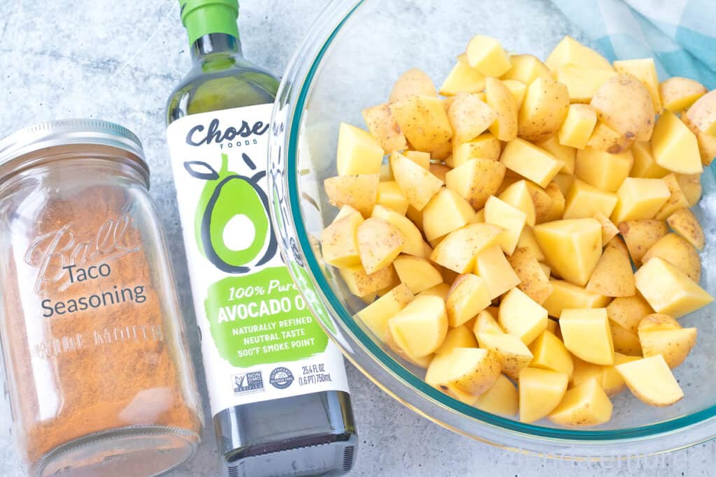 Ingredients to make Taco Potatoes on a concrete countertop — potato chunks, avocado oil and homemade taco seasoning.