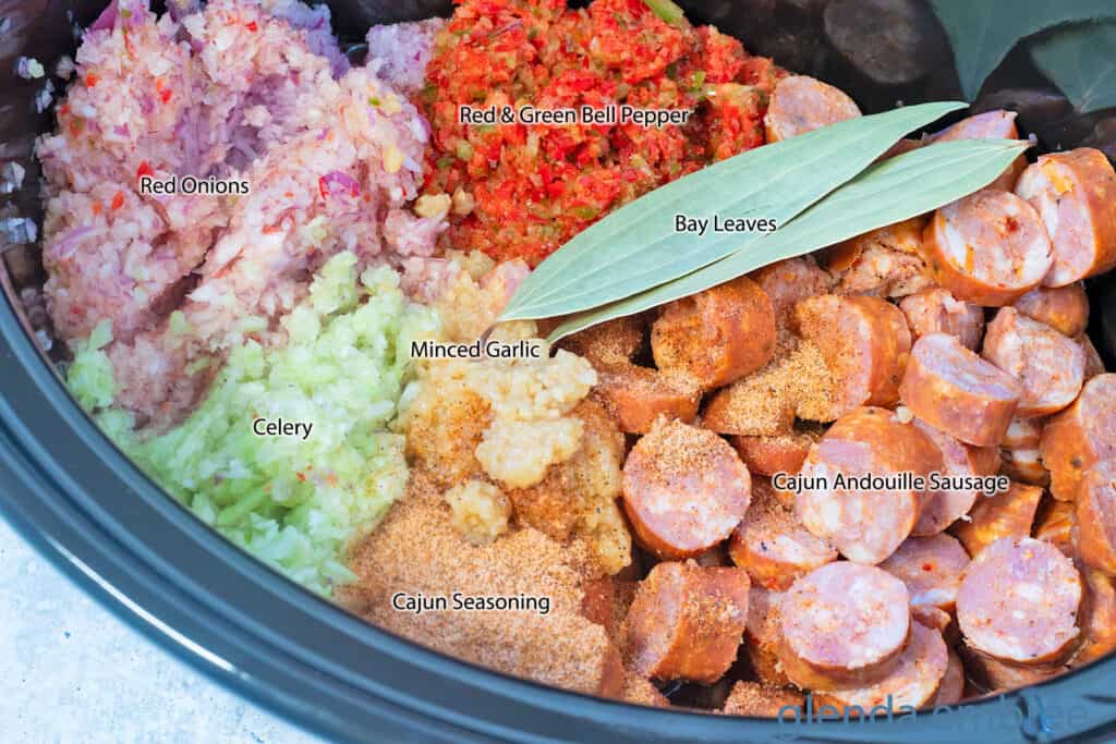 Andouille Sausage, Minced Garlic, Minced Red Onion, Minced red and green bell pepper, minced celery, Cajun seasoning, bay leaves and water in a crock pot ready to be cooked.