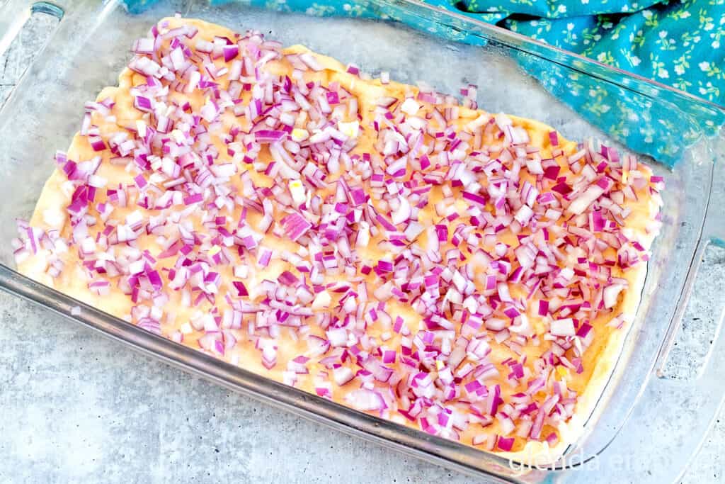 Adding the onion layer in a clear glass baking dish as Chicken Broccoli Cheese Casserole is assembled.