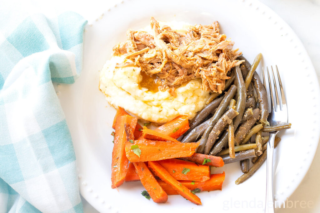 Slow Cooker Pork Roast over mashed potatoes on a white plate with green beans and carrots.